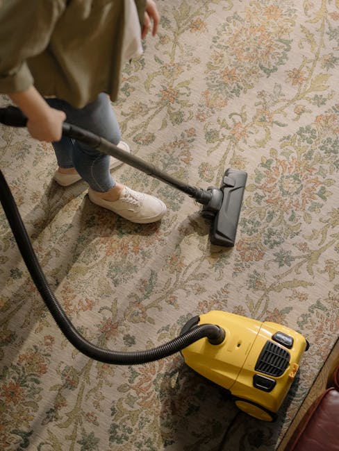 A person kneeling on a patterned indoor carpet, preparing a yellow and black vacuum cleaner for surface cleaning. The individual is wearing a beige jacket and blue jeans, and the vacuum's hose is exte