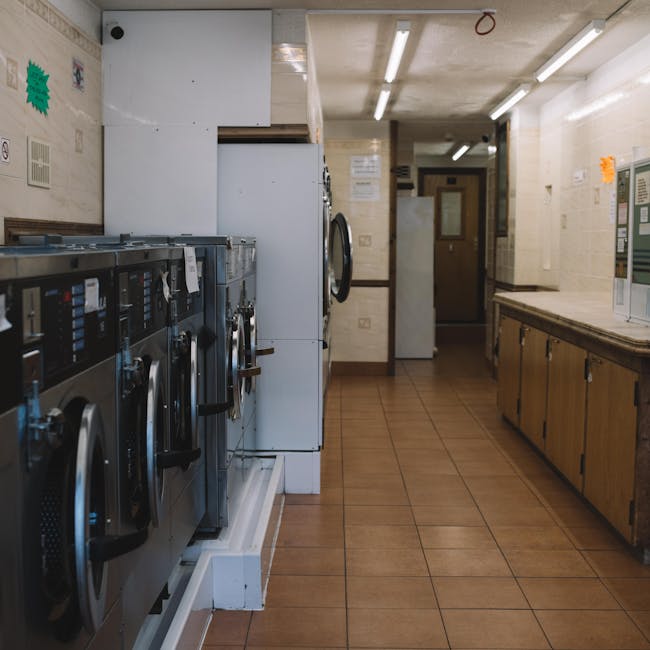 Interior view of a commercial laundry room or cleaning station with a row of stainless steel washing machines on the left, each with a digital control panel and door, situated on a white platform. To 