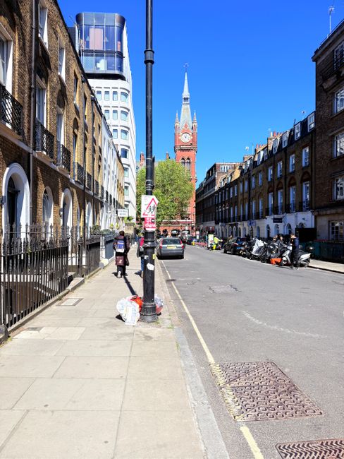 Two cleaning workers in bright orange and red high-visibility vests and pants are operating cleaning equipment on a city sidewalk in Finsbury Park, with one pushing a cart containing cleaning supplies